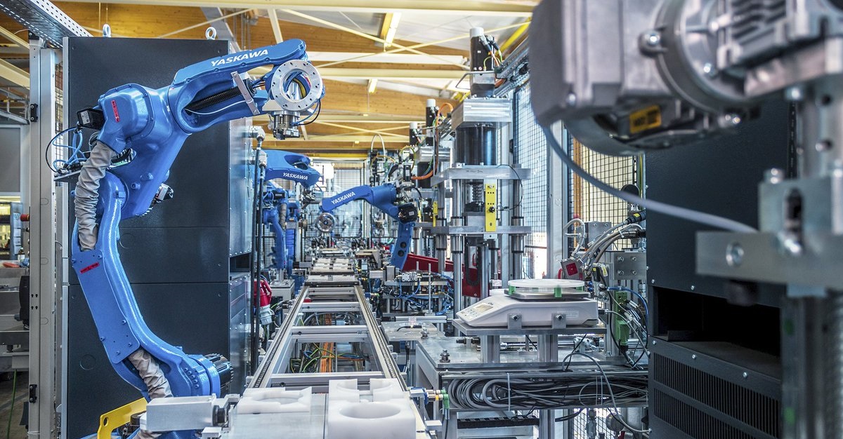 Automatiseringsproductielijn met Yaskawa Motoman robots die nauwkeurige bewerkingen uitvoeren in een industriële omgeving. De blauwe robotarmen zijn bezig met assemblage en productie, omringd door moderne technische apparatuur en componenten. Systeem geprogrammeerd door Robot Engineering.